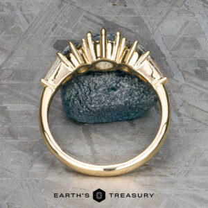The "Concordia" ring in 14k yellow gold, featuring a 0.94-carat Montana sapphire, is displayed upright against a rough bluish stone with a textured gray background.