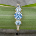 The "Concordia" ring in 14k yellow gold features a 0.94-carat Montana sapphire center, flanked by two round blue stones and two tapered clear stones, displayed vertically on a green stem with "Earth's Treasury" branding below.