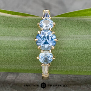 The "Concordia" ring in 14k yellow gold features a 0.94-carat Montana sapphire center, flanked by two round blue stones and two tapered clear stones, displayed vertically on a green stem with "Earth's Treasury" branding below.