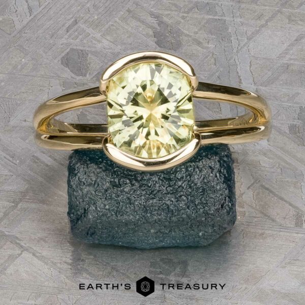 The "Madi" Ring in 14k yellow gold with 2.56-Carat Montana Sapphire The "Madi" Ring in 14k yellow gold with a 2.56-carat Montana sapphire stands upright on a rough blue-green stone, set against a geometric background with "Earth's Treasury" written below.