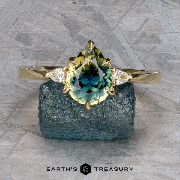 The "Sylph" ring in 14k yellow gold with 1.62-carat Australian sapphire The "Sylph" ring in 14k yellow gold features a 1.62-carat pear-shaped Australian sapphire and two small diamonds, shown on a rough blue stone with the "Earth's Treasury" logo below.