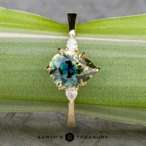 The "Sylph" ring in 14k yellow gold features a 1.62-carat round Australian sapphire in teal and yellow hues with two small pear-shaped side stones, showcased on a green leaf.