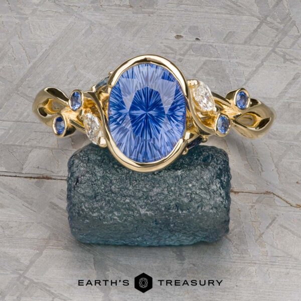 The "Cassia" ring in 18k yellow gold with 2.55-carat Madagascar sapphire The "Cassia" ring in 18k yellow gold features a 2.55-carat Madagascar sapphire, accented by smaller blue and marquise white stones, elegantly arranged on a rough stone surface.