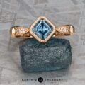 The "Myra" ring in 20k pink gold, featuring a 0.81-carat Montana sapphire, is displayed atop a rough blue stone on a textured gray background.