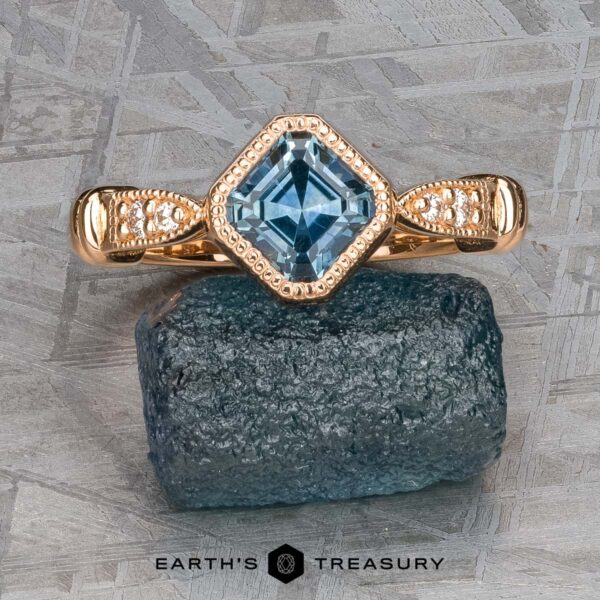 The "Myra" ring in 20k pink gold with 0.81-carat Montana sapphire The "Myra" ring in 20k pink gold, featuring a 0.81-carat Montana sapphire, is displayed atop a rough blue stone on a textured gray background.