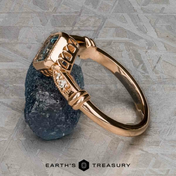 The "Myra" ring in 20k pink gold with 0.81-carat Montana sapphire The "Myra" ring in 20k pink gold with a 0.81-carat Montana sapphire is displayed upright against a rough dark stone on a textured surface, with the Earth's Treasury logo at the bottom.