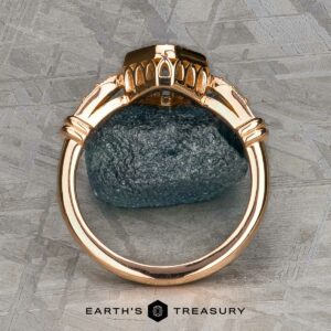 The "Myra" ring in 20k pink gold with a 0.81-carat Montana sapphire is displayed upright on a dark stone, set against a textured metallic background. The Earth's Treasury logo appears at the bottom.