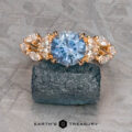 The "Yulania" ring in 20k pink gold with a 1.69-carat round Montana sapphire and clear marquise side stones is displayed on rough stone with the "Earth's Treasury" logo below.