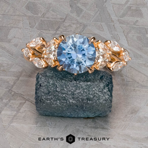 The "Yulania" ring in 20k pink gold with 1.69-Carat Montana Sapphire The "Yulania" ring in 20k pink gold with a 1.69-carat round Montana sapphire and clear marquise side stones is displayed on rough stone with the "Earth's Treasury" logo below.