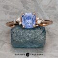 The "Iris" ring in 14k rose gold, set with a 1.14-carat Ceylon sapphire, is displayed on a rough teal mineral atop a textured gray surface.
