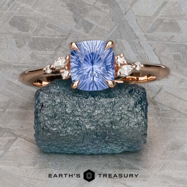 The "Iris" ring in 14k rose gold with 1.14-carat Ceylon sapphire The "Iris" ring in 14k rose gold, set with a 1.14-carat Ceylon sapphire, is displayed on a rough teal mineral atop a textured gray surface.