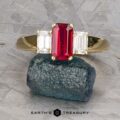 The "Olympia" ring in 18k yellow gold features a central 1.18-carat rectangular ruby flanked by two smaller clear gemstones, displayed on a rough stone.