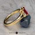 The "Olympia" ring in 18k yellow gold, featuring a 1.18-carat ruby and clear stone setting, is displayed on a gray textured surface next to a rough black stone.