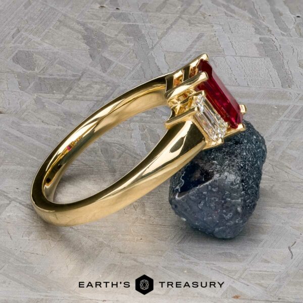 The "Olympia" ring in 18k yellow gold with 1.18-carat ruby The "Olympia" ring in 18k yellow gold, featuring a 1.18-carat ruby and clear stone setting, is displayed on a gray textured surface next to a rough black stone.