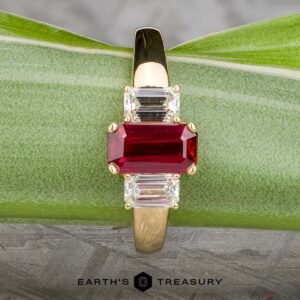 The "Olympia" ring in 18k yellow gold features a 1.18-carat rectangular ruby flanked by two smaller clear gemstones, set on a green leaf surface.