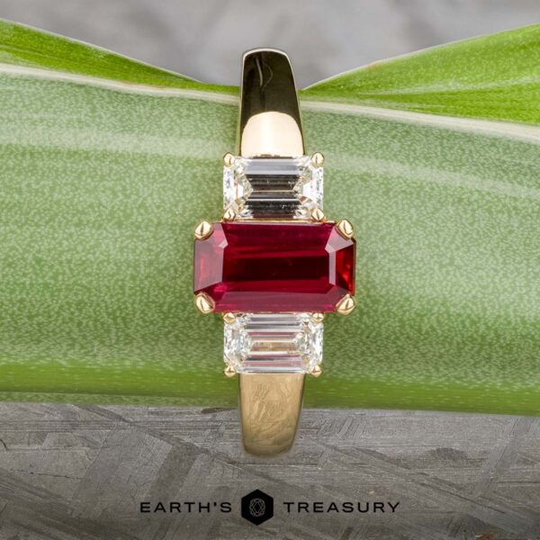 The "Olympia" ring in 18k yellow gold with 1.18-carat ruby The "Olympia" ring in 18k yellow gold features a 1.18-carat rectangular ruby flanked by two smaller clear gemstones, set on a green leaf surface.
