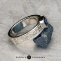 The "Kittley" ring in platinum with a 0.80-carat Montana Sapphire, adorned with engraved floral patterns, rests on rough blue mineral. "Earth's Treasury" logo is displayed at the bottom.