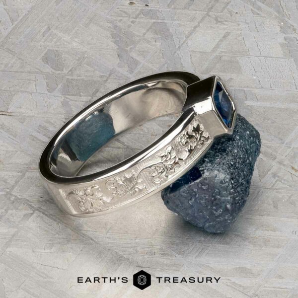 The "Kittley" ring in platinum with 0.80-carat Montana Sapphire The "Kittley" ring in platinum with a 0.80-carat Montana Sapphire, adorned with engraved floral patterns, rests on rough blue mineral. "Earth's Treasury" logo is displayed at the bottom.