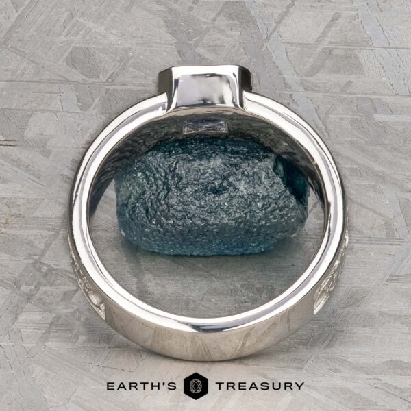 The "Kittley" ring in platinum with 0.80-carat Montana Sapphire The "Kittley" ring in platinum with a 0.80-carat Montana Sapphire rests on a textured gray surface over a rough blue gemstone. "EARTH'S TREASURY" below underscores the unique jewelry and fashion elegance of this piece.