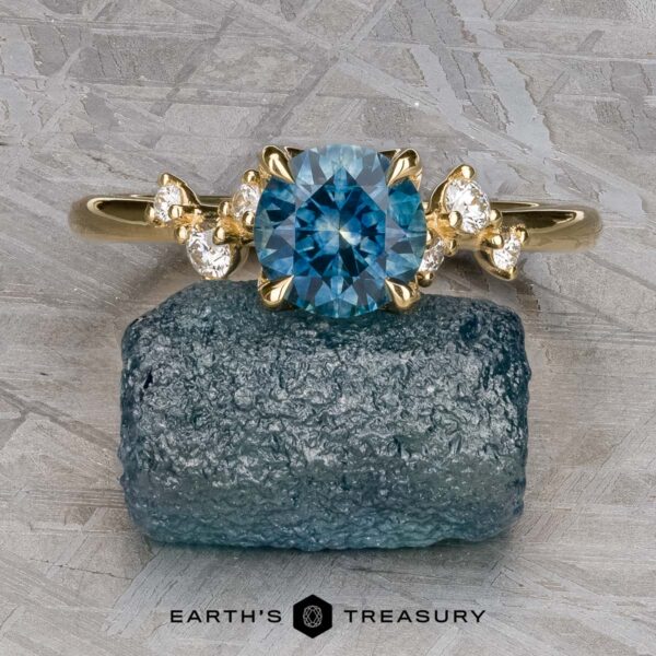 The "Poppy" ring in 18k yellow gold with 0.98-Carat Montana Sapphire The "Poppy" ring in 18k yellow gold showcases a 0.98-carat Montana sapphire with clear accent stones, displayed on rough blue crystal over a textured gray surface.