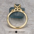 The "Poppy" ring in 18k yellow gold with a 0.98-carat Montana Sapphire stands upright against a blue stone, with the "Earth's Treasury" logo below on a textured background.