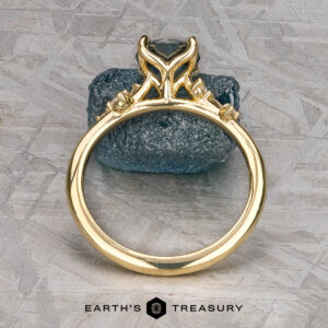 The "Poppy" ring in 18k yellow gold with a 0.98-carat Montana Sapphire stands upright against a blue stone, with the "Earth's Treasury" logo below on a textured background.