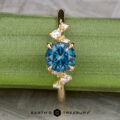 The "Poppy" ring in 18k yellow gold features a 0.98-carat round Montana Sapphire at the center, accented by small white stones, and is displayed on a green surface with "Earth's Treasury" branding below.