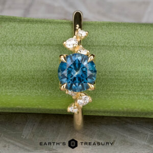 The "Poppy" ring in 18k yellow gold features a 0.98-carat round Montana Sapphire at the center, accented by small white stones, and is displayed on a green surface with "Earth's Treasury" branding below.