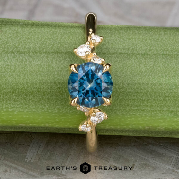 The "Poppy" ring in 18k yellow gold with 0.98-Carat Montana Sapphire The "Poppy" ring in 18k yellow gold features a 0.98-carat round Montana Sapphire at the center, accented by small white stones, and is displayed on a green surface with "Earth's Treasury" branding below.