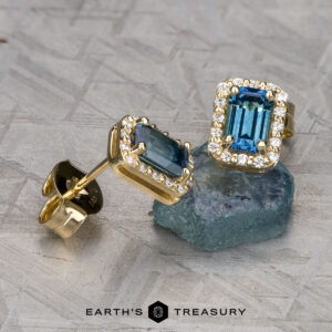Presenting the EA-MS-525: stunning gold stud earrings with rectangular blue gemstones, accented by small clear stones and showcased on a textured surface.