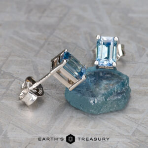 Product EA-MS-437, featuring silver earrings with rectangular blue gemstones, elegantly displayed on a raw blue crystal and set against a textured gray surface.
