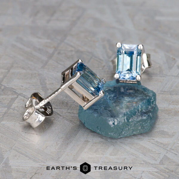 Product EA-MS-437, featuring silver earrings with rectangular blue gemstones, elegantly displayed on a raw blue crystal and set against a textured gray surface.