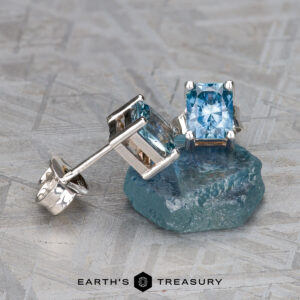 Two EA-MS-439 aquamarine earrings displayed on a textured gray surface, with one earring placed atop a rough blue stone. The earrings feature silver settings and square-cut gemstones.