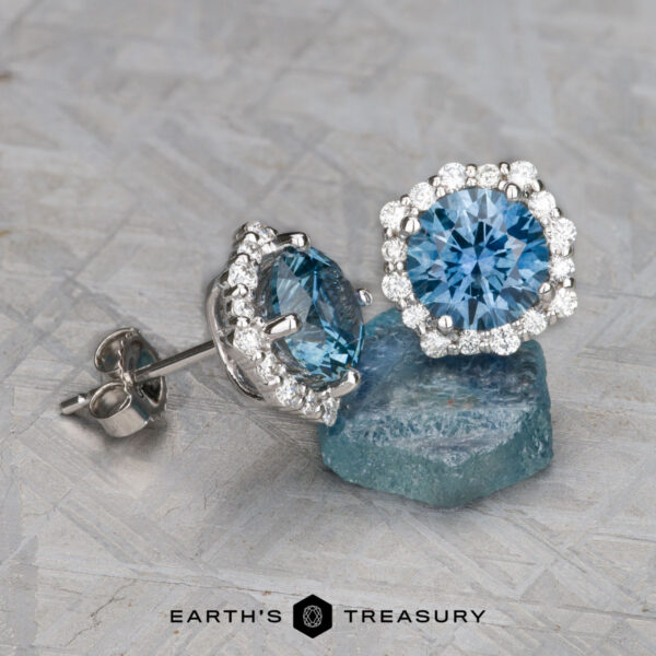 EA-MS-522 Two EA-MS-522 silver earrings with blue gemstones, surrounded by small diamonds, placed on a light textured surface next to a blue stone.