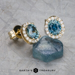 The EA-MS-523 earrings, featuring blue gemstones and diamond accents, are showcased alongside a rough blue stone on a textured surface. Text at the bottom reads "Earth's Treasury.