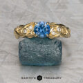 The "Viney" ring in 14k yellow gold with a 0.76-carat Montana sapphire, leaf and diamond accents is displayed on a rough blue mineral block against a geometric patterned background.