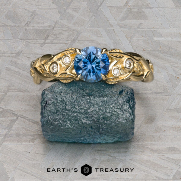 The "Viney" ring in 14K yellow gold with 0.76-carat Montana sapphire The "Viney" ring in 14k yellow gold with a 0.76-carat Montana sapphire, leaf and diamond accents is displayed on a rough blue mineral block against a geometric patterned background.