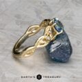 The "Viney" ring in 14k yellow gold features a 0.76-carat Montana sapphire and three diamonds, shown beside a rough blue mineral on a textured gray surface. Logo: "Earth's Treasury.