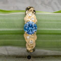The "Viney" ring in 14k yellow gold features a 0.76-carat round Montana sapphire at the center, accented by three small clear stones, and is displayed elegantly on a green striped leaf.