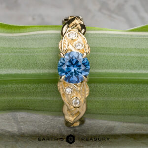 The "Viney" ring in 14k yellow gold features a 0.76-carat round Montana sapphire at the center, accented by three small clear stones, and is displayed elegantly on a green striped leaf.