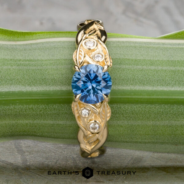 The "Viney" ring in 14K yellow gold with 0.76-carat Montana sapphire The "Viney" ring in 14k yellow gold features a 0.76-carat round Montana sapphire at the center, accented by three small clear stones, and is displayed elegantly on a green striped leaf.