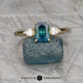 The "Nymph" ring in 14K yellow gold showcases a 1.25-carat Montana sapphire, flanked by two clear stones, and is displayed on rough blue crystal against a textured gray background for a striking presentation.