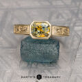 The "Kittley" ring in 14K yellow gold features an engraved band and a 1.45-carat octagonal Montana sapphire, set on a rough blue mineral and displayed on a textured gray surface.