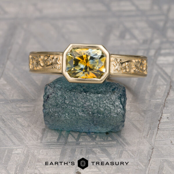 The "Kittley" ring in 14K yellow gold with 1.45-carat Montana sapphire The "Kittley" ring in 14K yellow gold features an engraved band and a 1.45-carat octagonal Montana sapphire, set on a rough blue mineral and displayed on a textured gray surface.