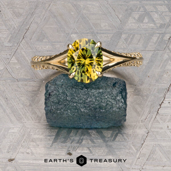 The "Antoinette" ring in 14k yellow gold with 1.69-carat Australian sapphire The "Antoinette" ring in 14k yellow gold, set with a 1.69-carat Australian sapphire, stands upright on a rough blue stone against a patterned backdrop, with the "EARTH'S TREASURY" logo below.