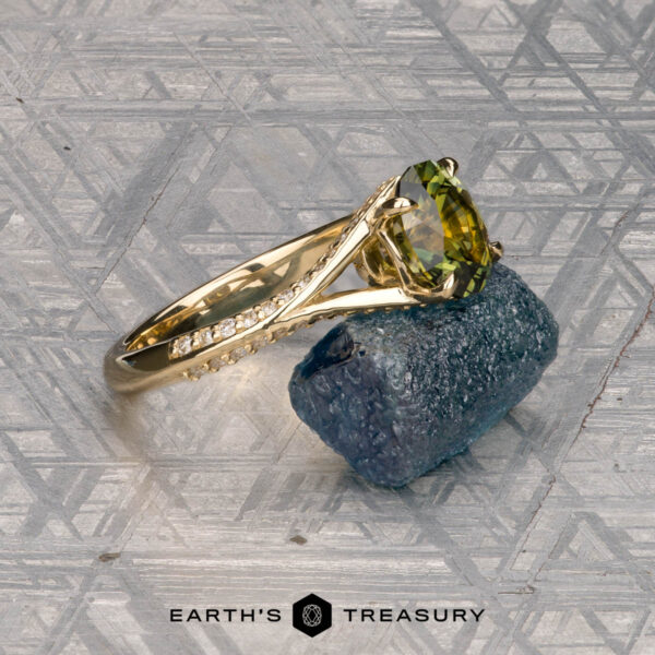 The "Antoinette" ring in 14k yellow gold with 1.69-carat Australian sapphire The "Antoinette" ring, crafted in 14k yellow gold with a 1.69-carat Australian sapphire, is showcased atop a rough blue crystal with a geometric patterned background.