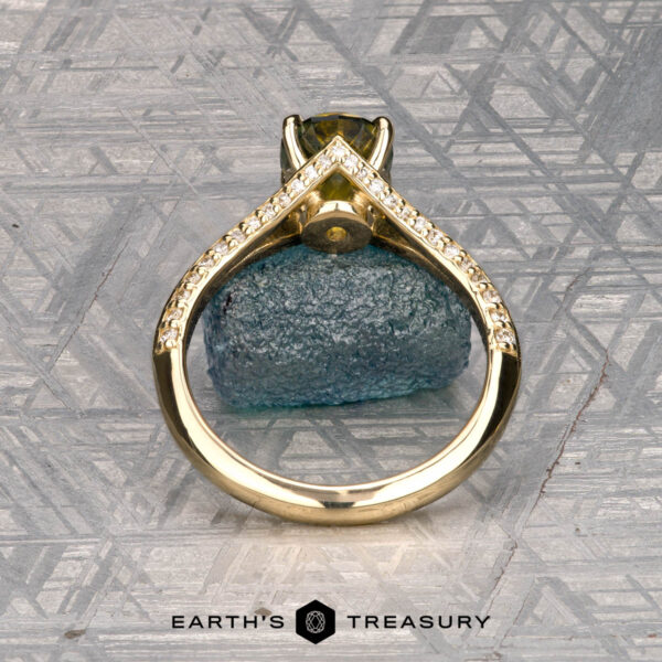 The "Antoinette" ring in 14k yellow gold with 1.69-carat Australian sapphire The "Antoinette" ring in 14k yellow gold with a 1.69-carat Australian sapphire stands upright before a rough blue mineral, with small diamonds and a geometric metallic background. "Earth's Treasury" appears at the bottom.