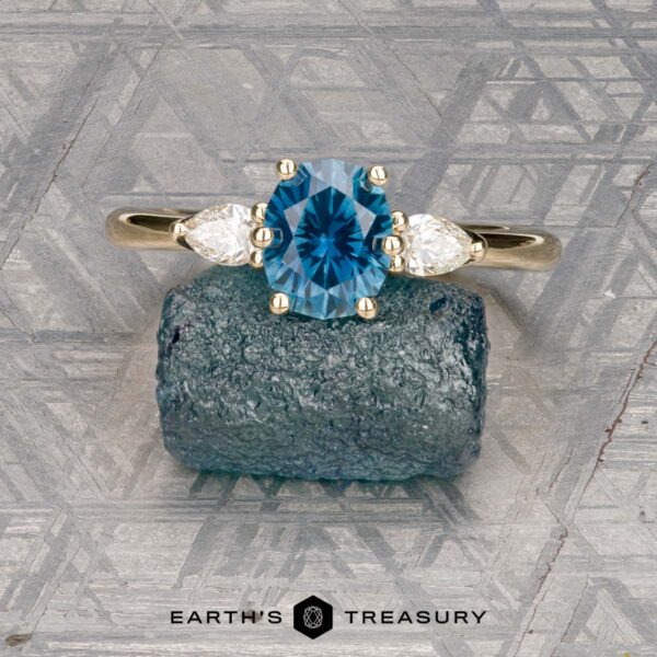 The "Nymph" ring in 14K yellow gold with 1.27-carat Montana sapphire The "Nymph" ring in 14K yellow gold features a 1.27-carat Montana sapphire accented by two pear-shaped clear stones, displayed on a rough blue crystal with the "Earth’s Treasury" logo—a striking three-stone ring for any occasion.