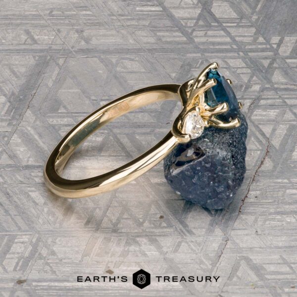 The "Nymph" ring in 14K yellow gold with 1.27-carat Montana sapphire The "Nymph" ring in 14K yellow gold features a 1.27-carat Montana sapphire accented by two pear-shaped clear stones, displayed on a rough blue crystal with the "Earth’s Treasury" logo—a striking three-stone ring for any occasion.