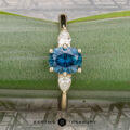 The "Nymph" ring in 14K yellow gold features a 1.27-carat Montana sapphire flanked by two pear-shaped clear stones, beautifully presented on a green leaf background for an enchanting, nature-inspired look.